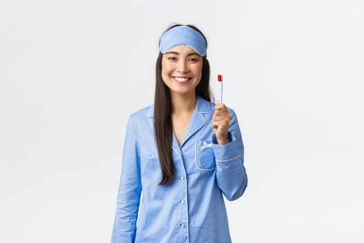 Hygiene, lifestyle and people at home concept. Cheerful smiling asian girl in pajamas and sleeping mask showing toothbrush and white perfect teeth, use whitening toothpaste, white background