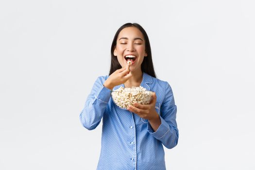 Home leisure, sleepover and slumber party concept. Smiling delighted asian girl enjoying eating favorite popcorn, wearing pajama on weekend, watching favorite movie, white background