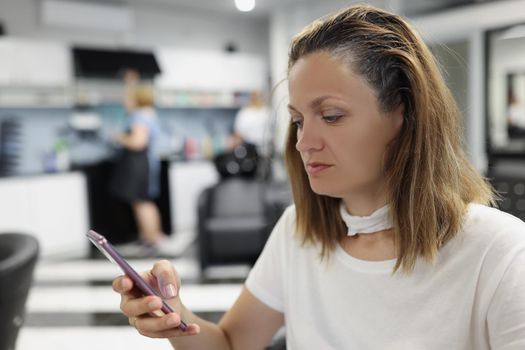 Woman client sit at hairdresser, woman use smartphone while waiting for hair to dye