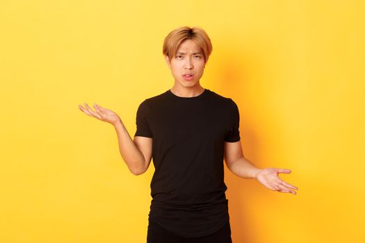 Portrait of confused and annoyed asian guy spread hands sideways, cant understand something, standing yellow background