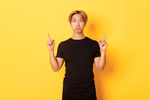 Portrait of sulking gloomy asian guy looking disappointed, pointing fingers up, yellow background