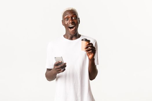 Portrait of excited and amazed blond african-american man, looking fascinated, using mobile phone and drinking coffee, standing white background.