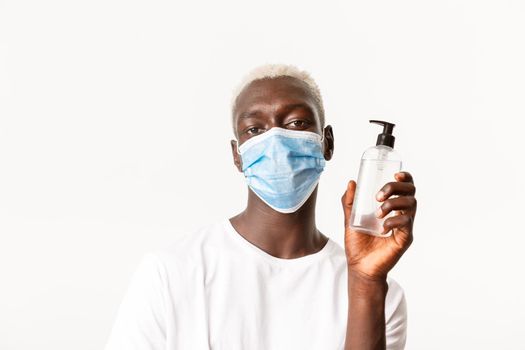 Portrait of stylish handsome african-american blond male in medical mask, showing hand sanitizer, advice using preventive measures during coronavirus pandemic, white background