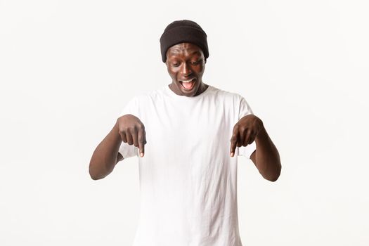 Portrait of excited, happy african-american man in beanie, yelling from amazement and pointing fingers down, looking at your logo cheerful, standing white background
