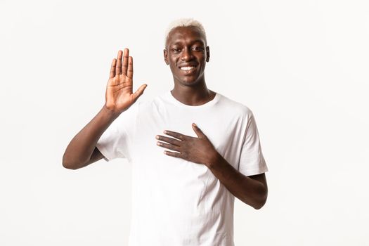 Portrait of honest attractive african-american blond guy, raising one hand and another on heart, making promise, smiling over white background
