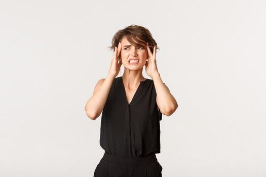 Young woman touching temples and grimacing from painful migraine. Girl complaining on headache, standing white background