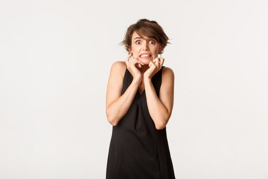 Image of scared timid girl in black dress, looking frightened and trembling from fear, standing over white background