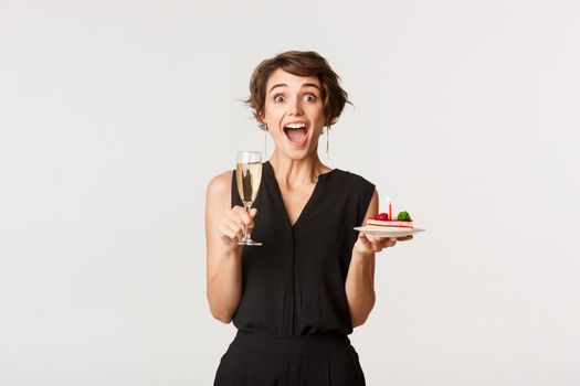 Image of young amazed woman attend birthday celebration party, holding glass of champagne and cake.