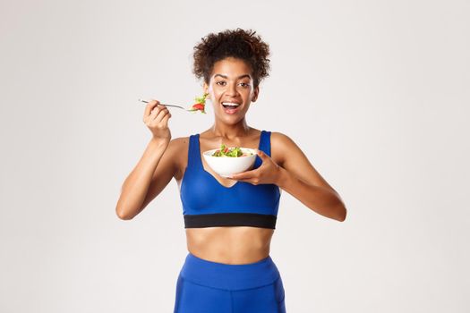 Healthy lifestyle and sport concept. Beautiful african-american fitness woman eating salad and smiling, standing in sport costume.