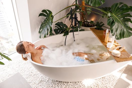 Relaxed young woman bathing with body care cosmetics on wooden shelf over modern white bubble filled bathtub at home