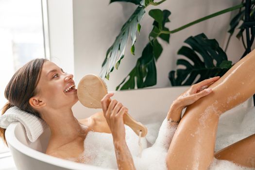 Cute young woman relaxing in bathtub and singing into a brush while spending time in bathroom decorated with tropical plant