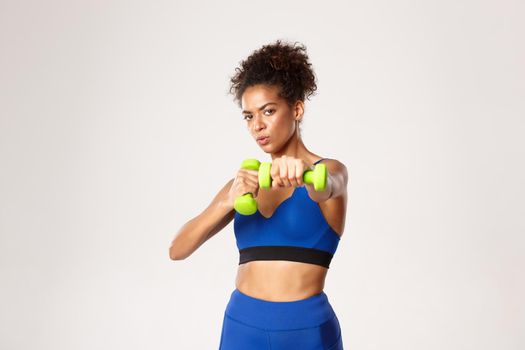 Concept of sport and workout. Determined african-american fitness woman, boxing, practice punches with dumbbells, standing against white background.