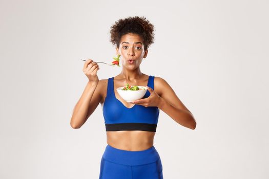 Healthy lifestyle and sport concept. Fit african-american fitness woman looking surprised while eating salad, standing over white background.