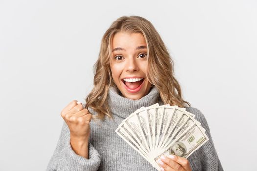 Close-up of successful and rich blond girl in grey sweater, holding money and looking like winner, standing over white background