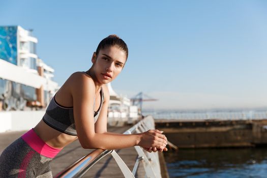 Close-up of attractive female athlete leaning on a handrail and looking at the sea, finish workout on the seaside promenade