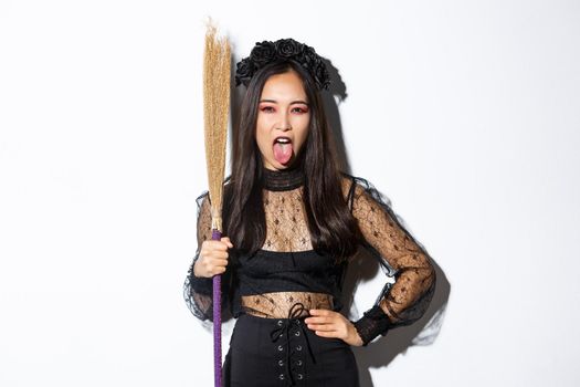 Carefree sassy asian woman enjoying halloween party, wearing witch costume and holding broom, showing tongue, standing over white background