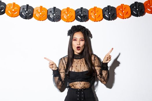 Joyful asian woman in gothic lace dress celebrating halloween, pointing fingers sideways, standing against white background with pumpkin party streamers
