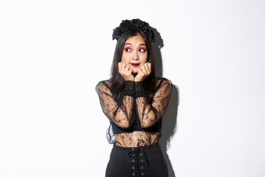 Silly beautiful asian girl in gothic lace dress with wreath looking dreamy left, have nostalgic thoughts, standing in halloween costume over white background