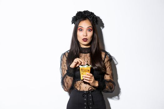 Image of surprised and fascinated asian woman in witch costume, holding sweets gathered during trick or treating, standing over white background