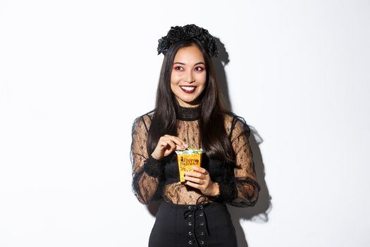 Smiling cute asian woman celebrating halloween, holding sweets and grinning happy, trick or treating in witch costume