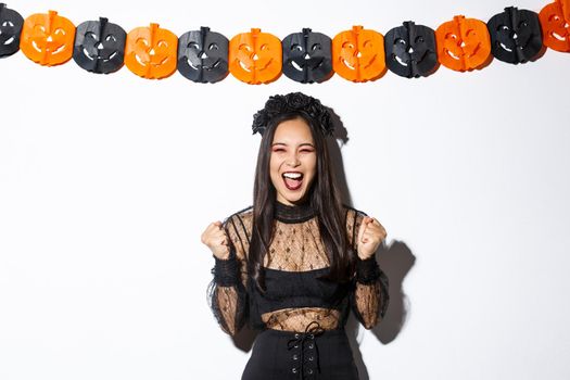 Happy asian woman enjoying halloween, wearing wicked witch costume and rejoicing against pumpkin streamers decoration