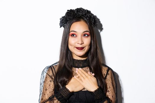 Close-up of nostalgic smiling asian woman looking at upper left corner dreamy, holding hands on heart, wearing gothic lace dress for halloween party