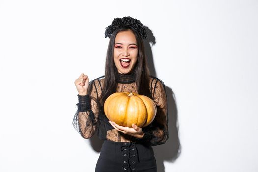 Image of excited beautiful asian woman celebrating halloween, wearing witch outfit and holding pumpkin, yelling with rejoice