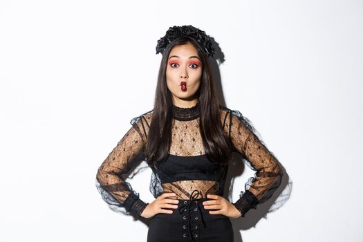 Portrait of silly attractive asian woman fooling around, showing funny faces at camera while wearing halloween costume, standing over white background