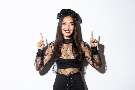 Beautiful asian woman in undead widow costume, wearing black lace dress for halloween, pointing fingers up and smiling pleased, looking at your logo, standing over white background