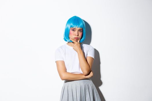 Portrait of troubled asian girl in blue wig pouting, looking complicated while pondering something, standing over white background