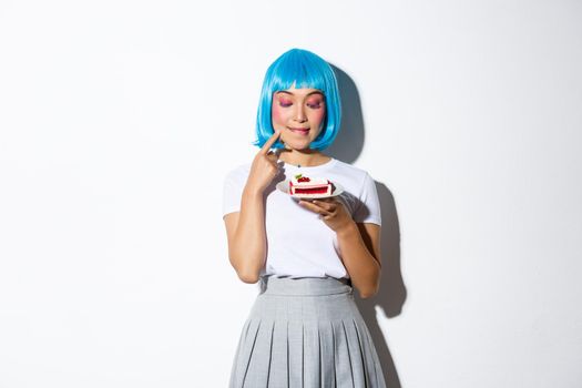 Image of coquettish fashion girl in blue wig looking with temptation at cake, standing over white background