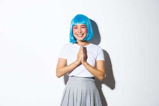 Portrait of lovely smiling asian girl in blue party wig, thanking for something, clasp hands together and looking grateful, standing over white background