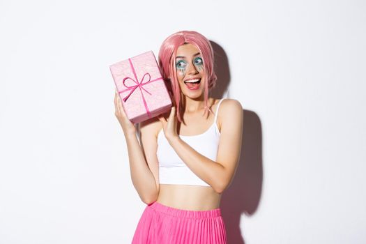 Image of beautiful girl in pink wig shaking box with birthday gift, wonder what inside wrapped box, standing over white background