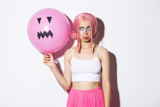 Moody cute girl in pink wig, pouting jealous or displeased, standing with jack-o-lantern balloon, celebrating halloween, standing over white background