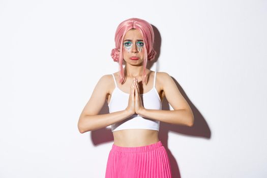 Portrait of miserable cute girl in pink anime wig asking for help, clasp hands together and begging for something, standing in halloween outfit over white background