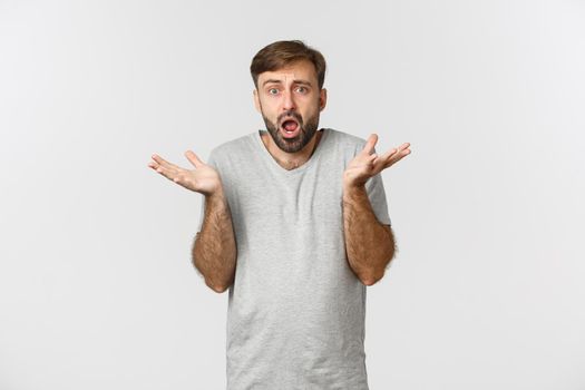 Portrait of confused and worried bearded guy, wearing casual t-shirt, looking at something complicated and shrugging, standing over white background