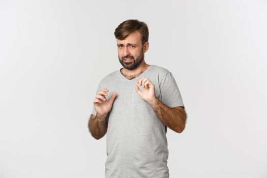 Portrait of timid and scared man in gray t-shirt, raising hands to defend himself, looking frightened, standing over white background