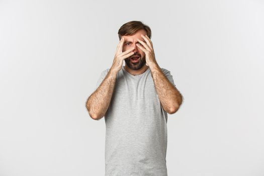 Portrait of embarrassed bearded man in gray t-shirt, cover eyes with hands but peeking through fingers scared to look, standing over white background