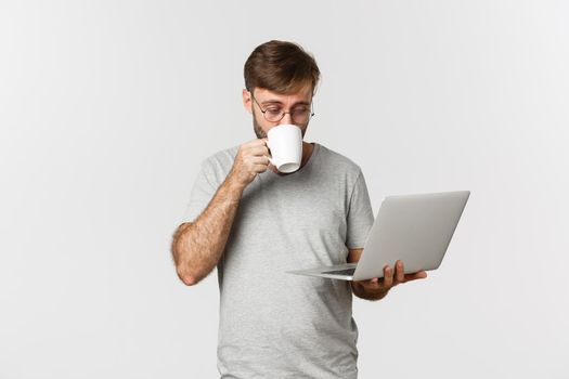 Image of male freelancer working with laptop, drinking coffee, standing in gray t-shirt and glasses over white background