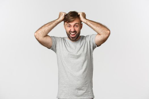 Portrait of distressed mad guy in gray t-shirt, ripping hair from anger and shouting, standing over white background