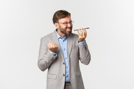 Pissed-off and angry businessman with beard, wearing grey suit and glasses, shouting at speakerphone with mad expression, standing over white background