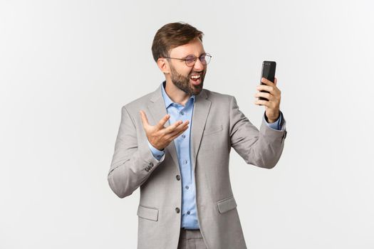 Portrait of businessman in grey suit and glasses, having video call, looking disappointed and confused at smartphone, having an argument, standing over white background.
