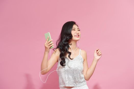 Portrait of charming joyful and emotive woman smiling from joy listening music in earphones holding smartphone posing against pink wall