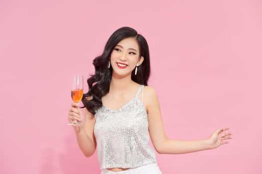 Good-looking asian girl in stylish attire standing in confident pose with wineglass on pink background. 