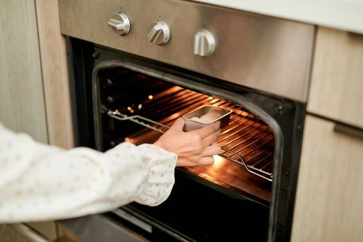woman wearing oven mitt putting baking sheet with raw cookies into modern oven in home kitchen