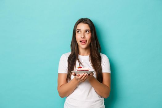 Image of cute brunette girl celebrating her birthday, licking lips as holding delicious b-day cake, making wish, standing over blue background