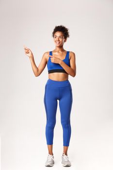 Full length of athletic african-american gitness girl in blue sportswear, pointing fingers left and looking pleased, recommend workout equipment, standing over white background.