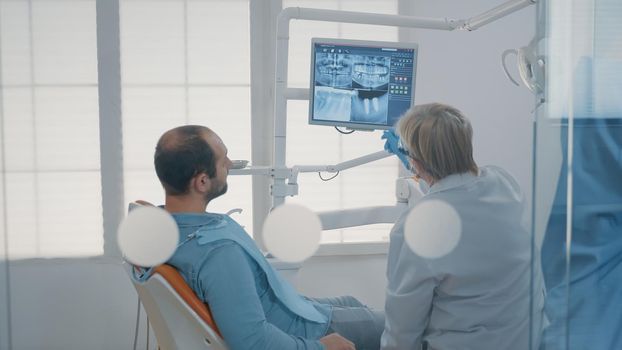 Mature dentist pointing at teeth radiography on monitor to explain dentititon diagnosis to patient with toothache. Dental expert showing x ray scan results to man with cavity pain.