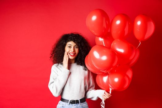 Holidays and celebration. Surprised birthday girl holding helium party balloons and looking touched at camera, being congratulated, red background