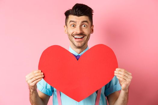 Valentines day concept. Romantic man falling in love, showing big red heart cutout and smiling, standing on pink background